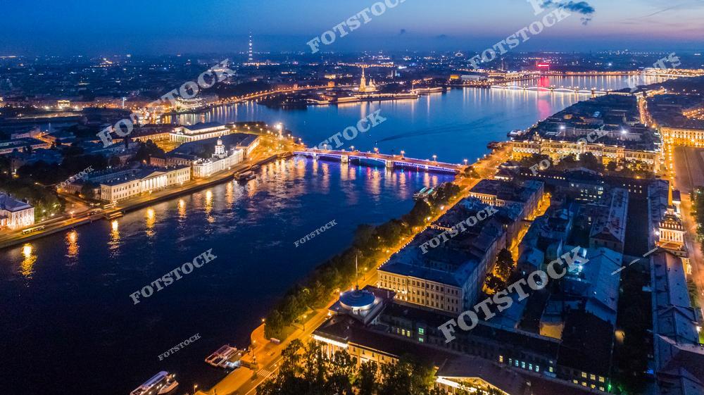 На фотографии изображён ночной Санкт-Петербург, подсвеченный огнями белых ночей. Видны река Нева, огни мостов и набережных, а также архитектурные достопримечательности города. Ярко выделяются стрелка Васильевского острова и Петропавловская крепость. Городская панорама с высоты передаёт атмосферу живого, ночного Петербурга.